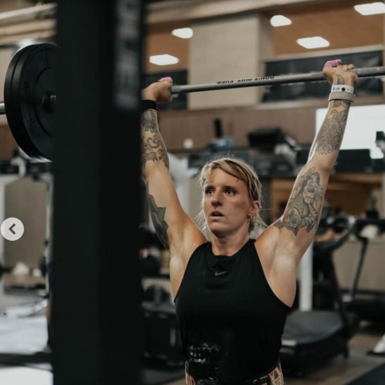 Female athlete pressing a loaded barbell overhead during an LT Games strength segment.