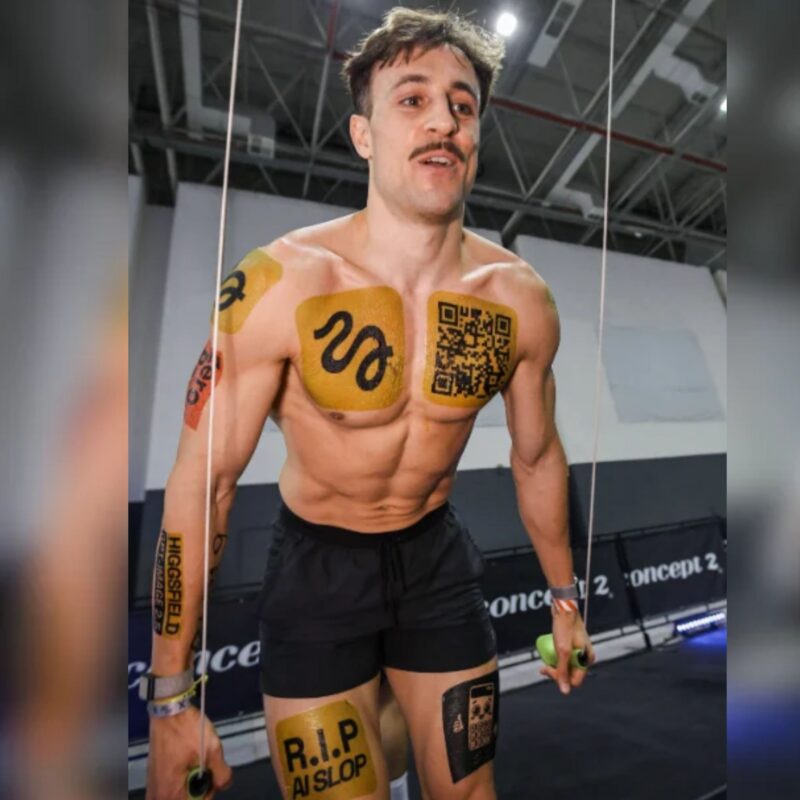 Marc Lou on the SkiErg at a HYROX race with gold sponsor logo tattoos on his chest, shoulder, arm and thighs